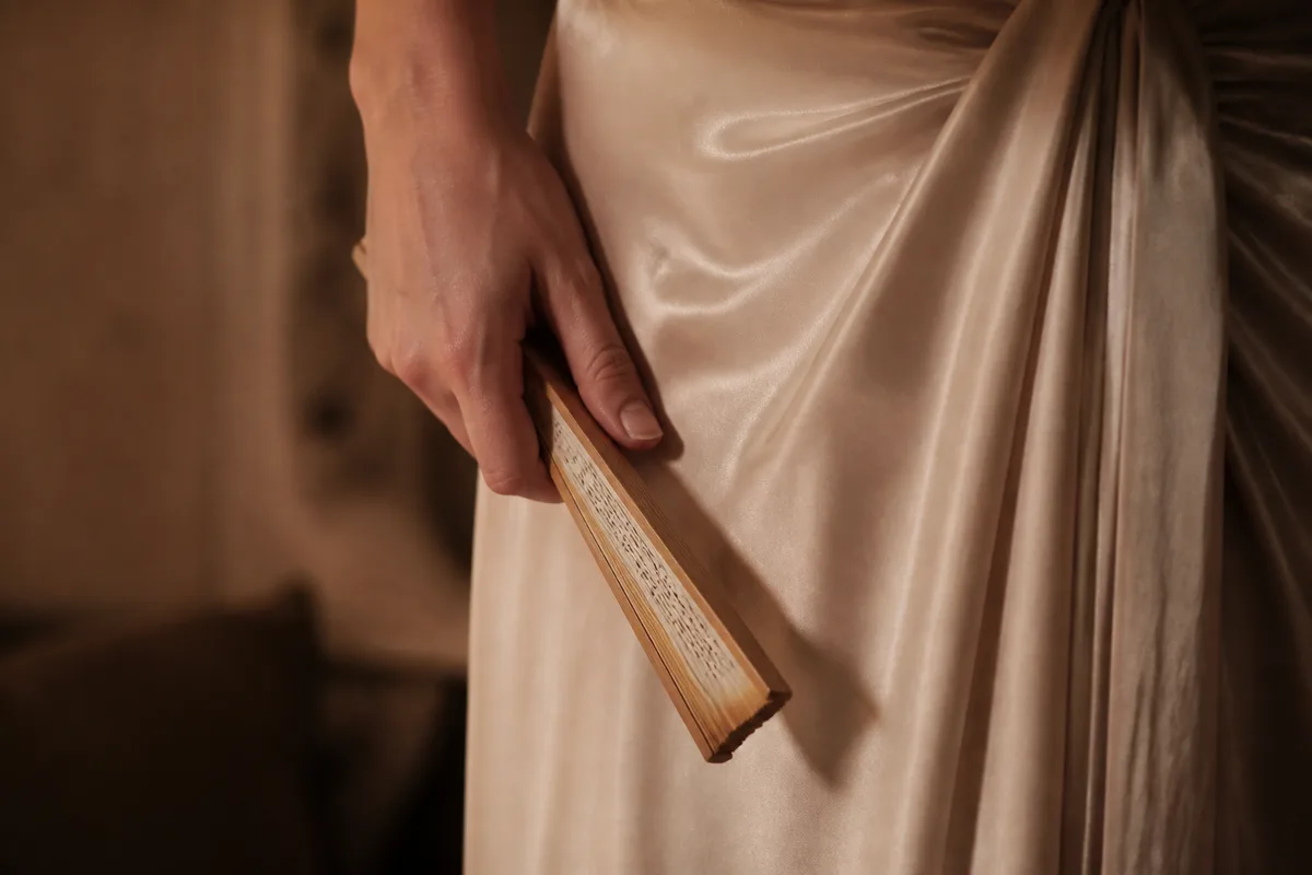 Détail d&rsquo;une robe de satin clair et d&rsquo;une main tenant un éventail fermé